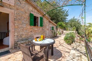 Un bâtiment en pierre avec une table ronde et des chaises en osier, un petit jardin avec des plantes et un ciel dégagé.