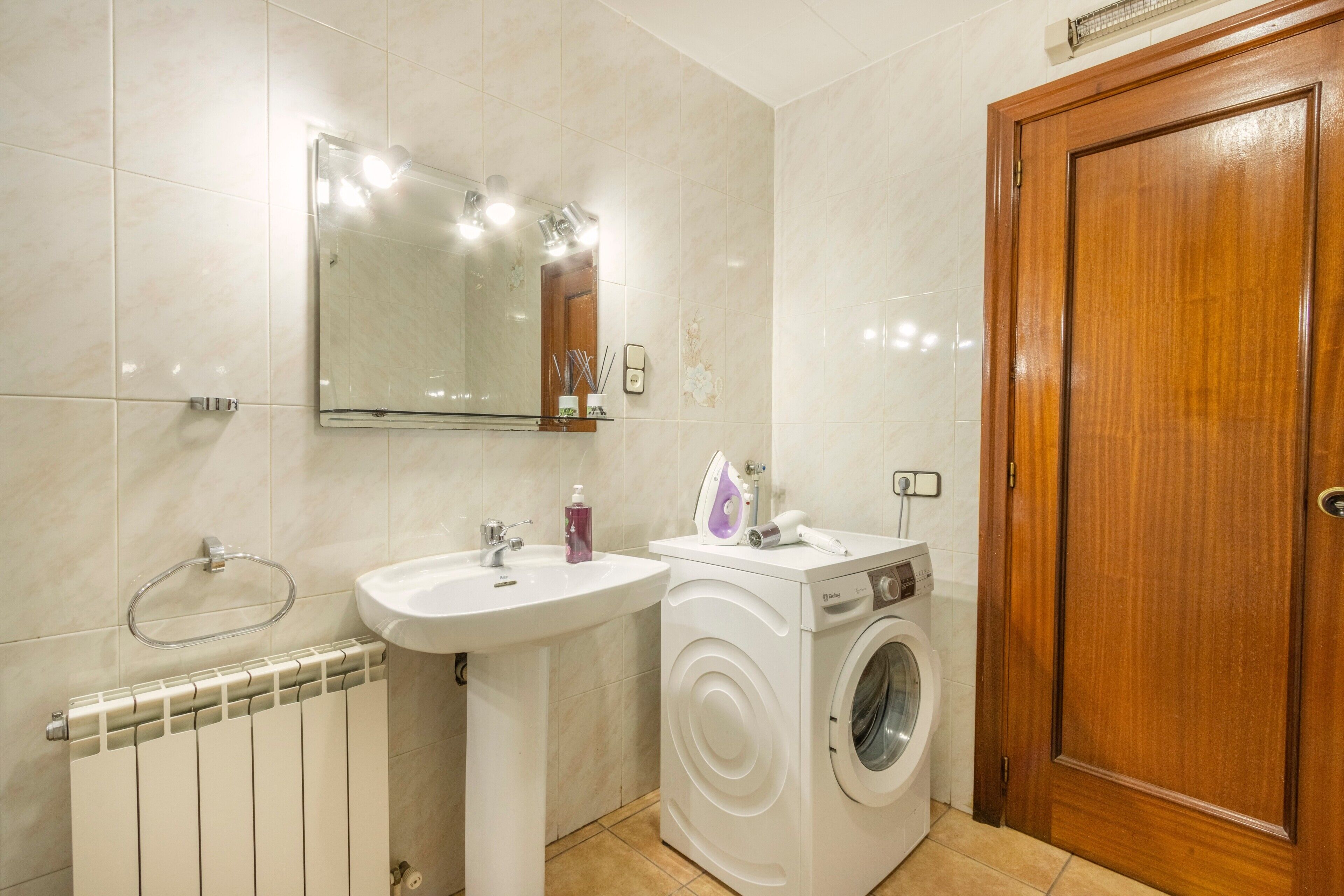 A bathroom with a sink, mirror, radiator, washing machine, and wooden door.