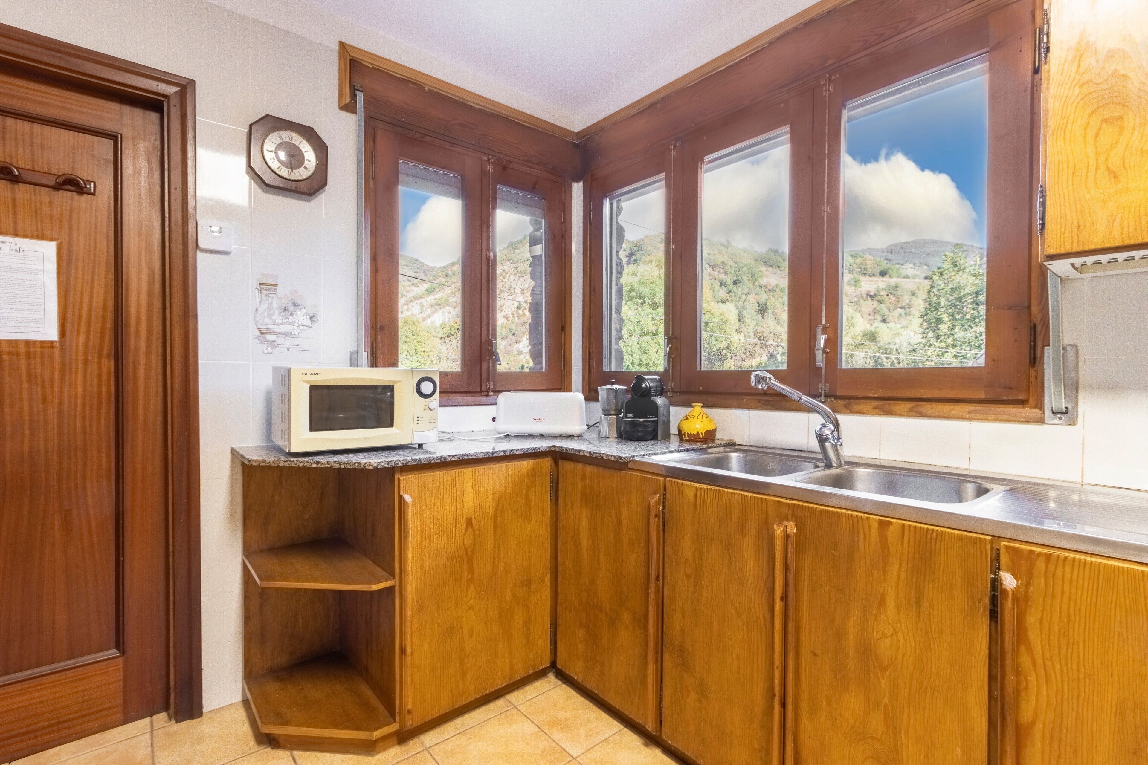 Una cocina con armarios de madera, microondas, tostadora, lavabo y ventanas amplias con vista a la montaña.