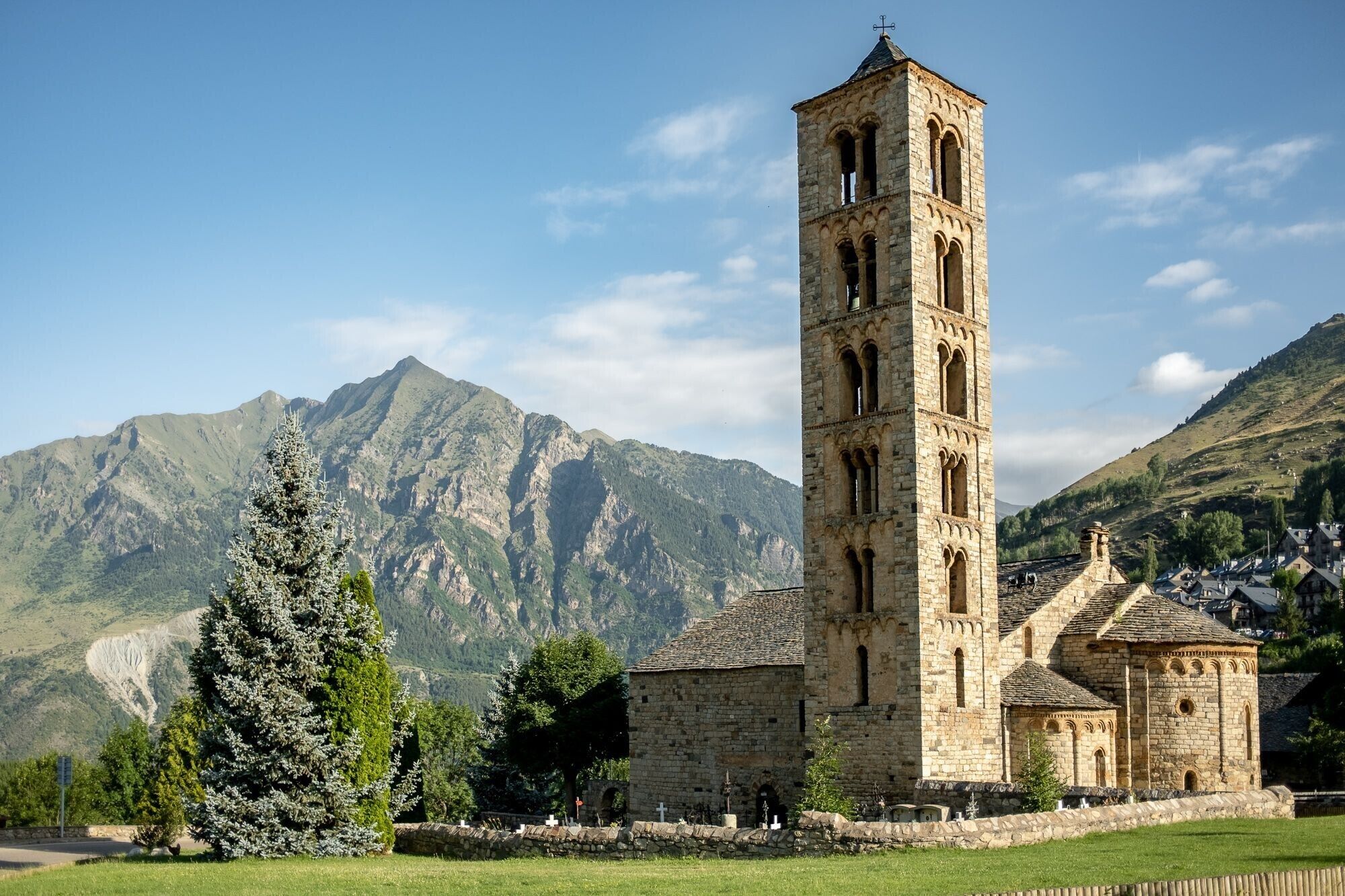 Una iglesia de piedra histórica con un campanario alto, rodeada de montañas y un pequeño pueblo.