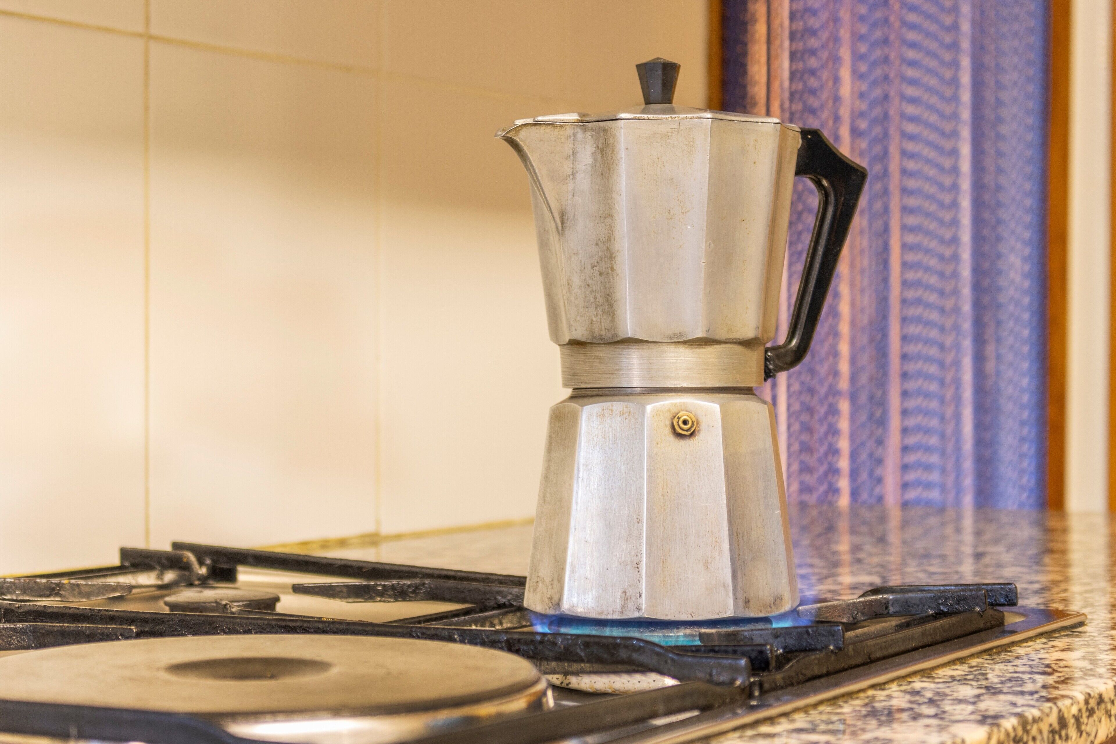 A stainless steel coffee maker on a gas stove with a tiled backsplash.