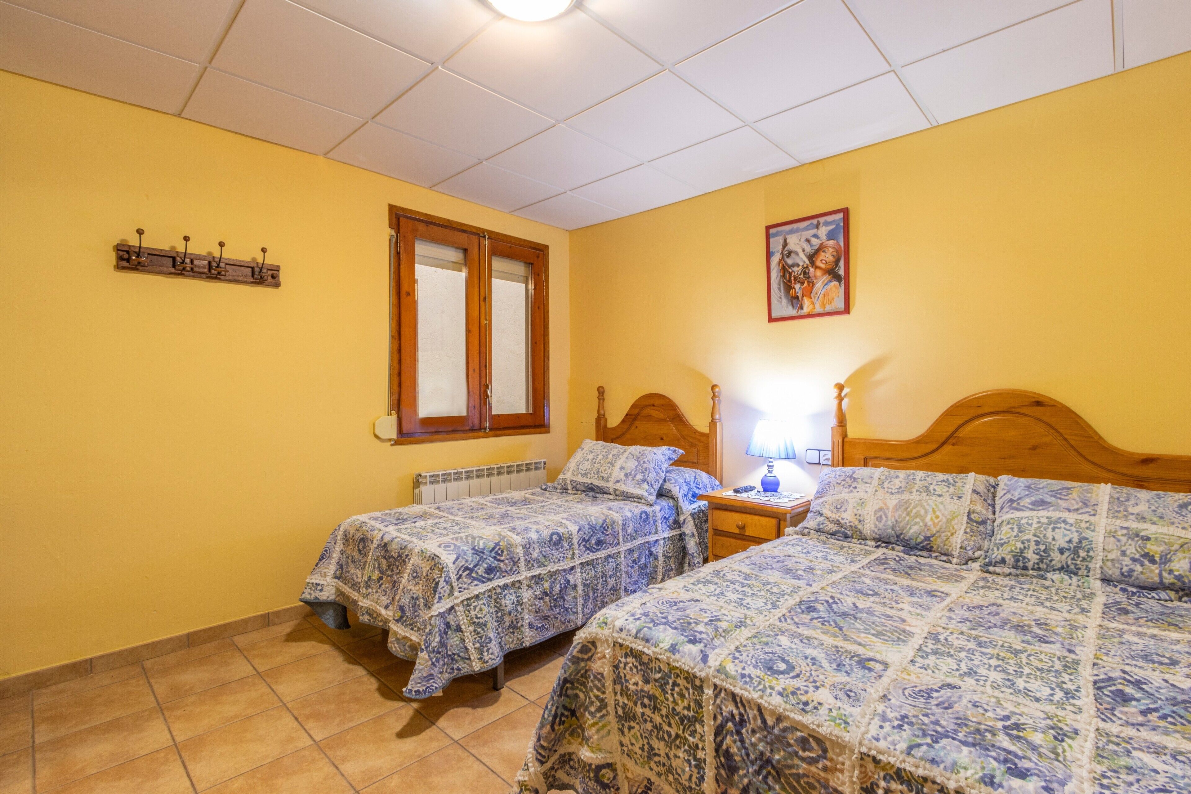 A bedroom with two beds, a window, a radiator, and a wall-mounted shelf.
