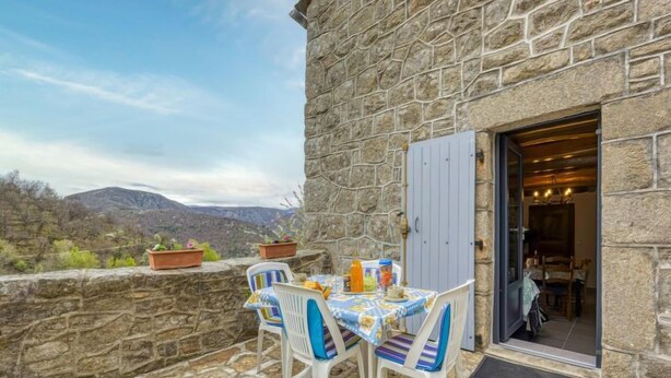 A stone building with a table and chairs on a terrace overlooking a mountainous landscape.