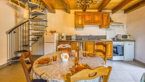 A dining area with a round table, chairs, a chandelier, wooden cabinets, a kitchen with appliances, and a spiral staircase.