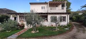 Una casa de dos pisos con un porche cubierto, un pequeño jardín con árboles y rocas, y un camino de entrada pavimentado.