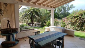 Un porxo cobert amb una taula i cadires de fusta, una prestatgeria per a la roba penjada, i vista a un jardí amb palmeres i arbustos florits.