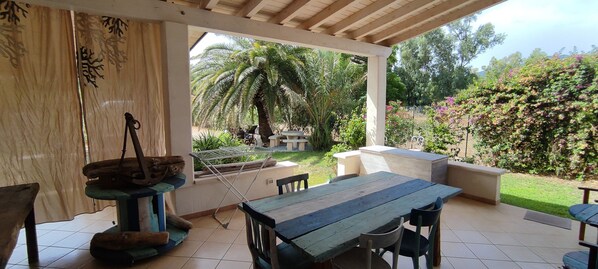 Un porche techado con una mesa y sillas de madera, un tendedero colgante y vistas a un jardín con palmeras y arbustos florecientes.
