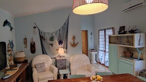 Un saló amb televisió, dues butaques blanques, una taula de fusta amb un plat de menjar, una decoració de xarxa penjant i elements decoratius marins a la paret.