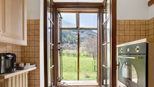 A kitchen with an open door leading to a view of a green landscape, a coffee maker, and a stove. - Apartment 'Casa Tua - Avise' with Mountain View, Private Terrace and Wi-Fi (Runaz)