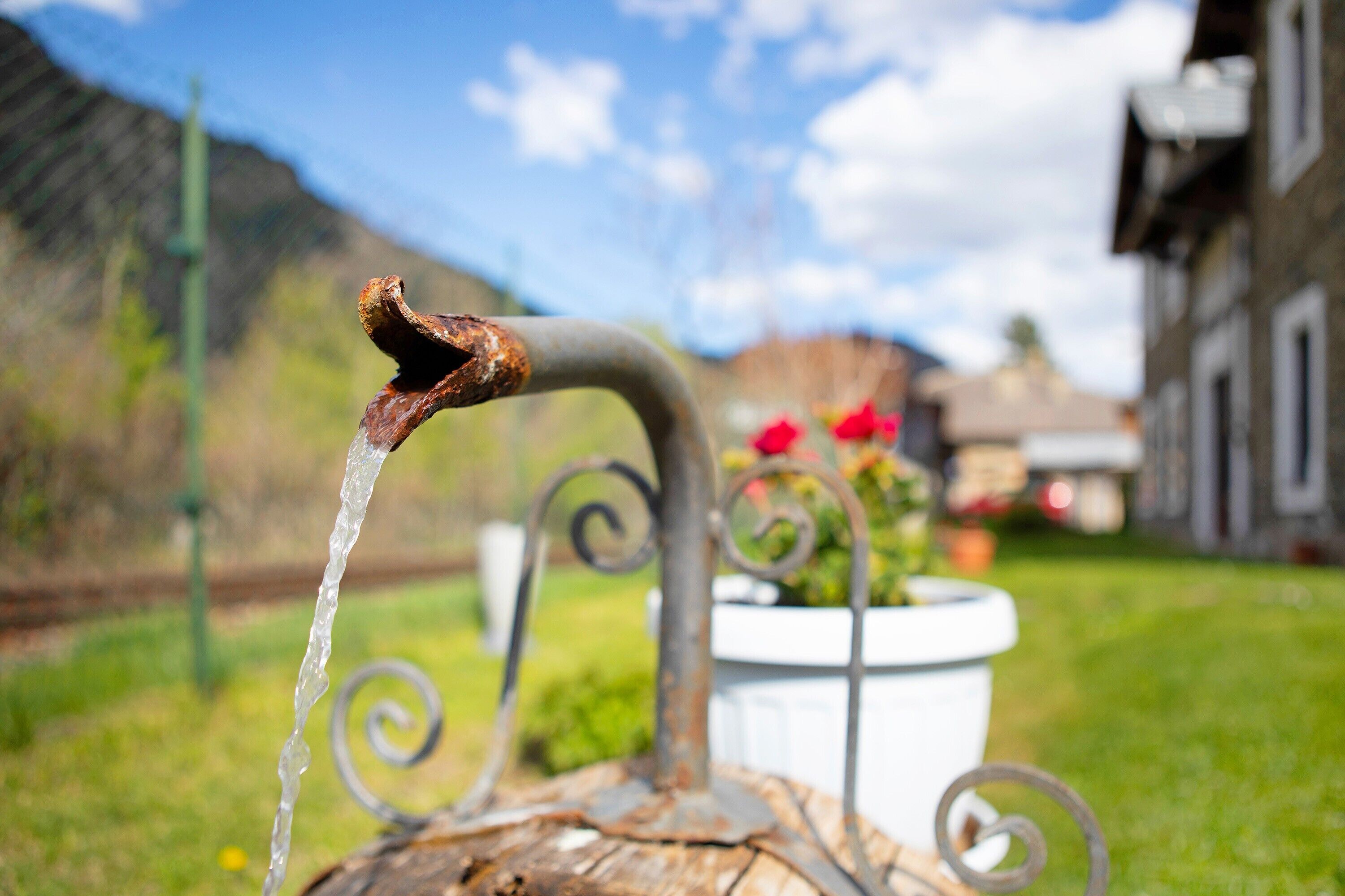 A rustic outdoor water spout with a metal design, a white planter with red flowers, and a building in the background.