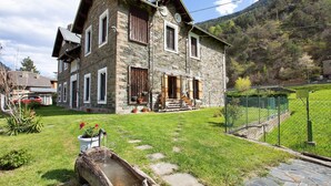 A stone house with a front porch, a small garden, and a mountain backdrop. - Apartment 'Casa Tua - Avise' with Mountain View, Private Terrace and Wi-Fi (Runaz)