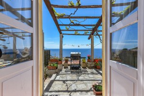 Ein gepflasterter Weg führt zu einer Holzw pergola mit Grill, Blumentöpfen und Blick auf das Meer.
