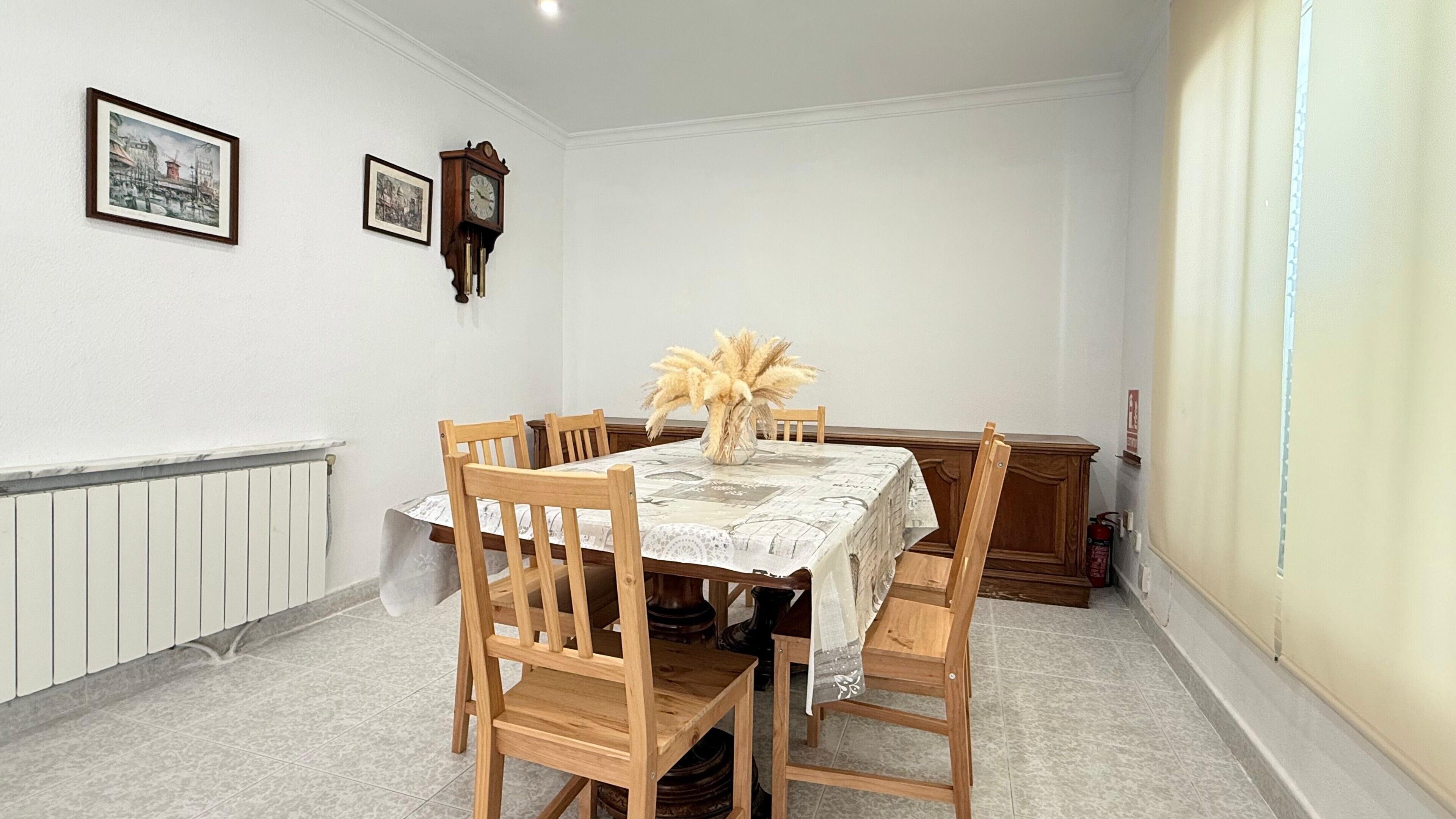 A dining room with a table set for six, wooden chairs, a vase with dried flowers, and two framed pictures on the wall.