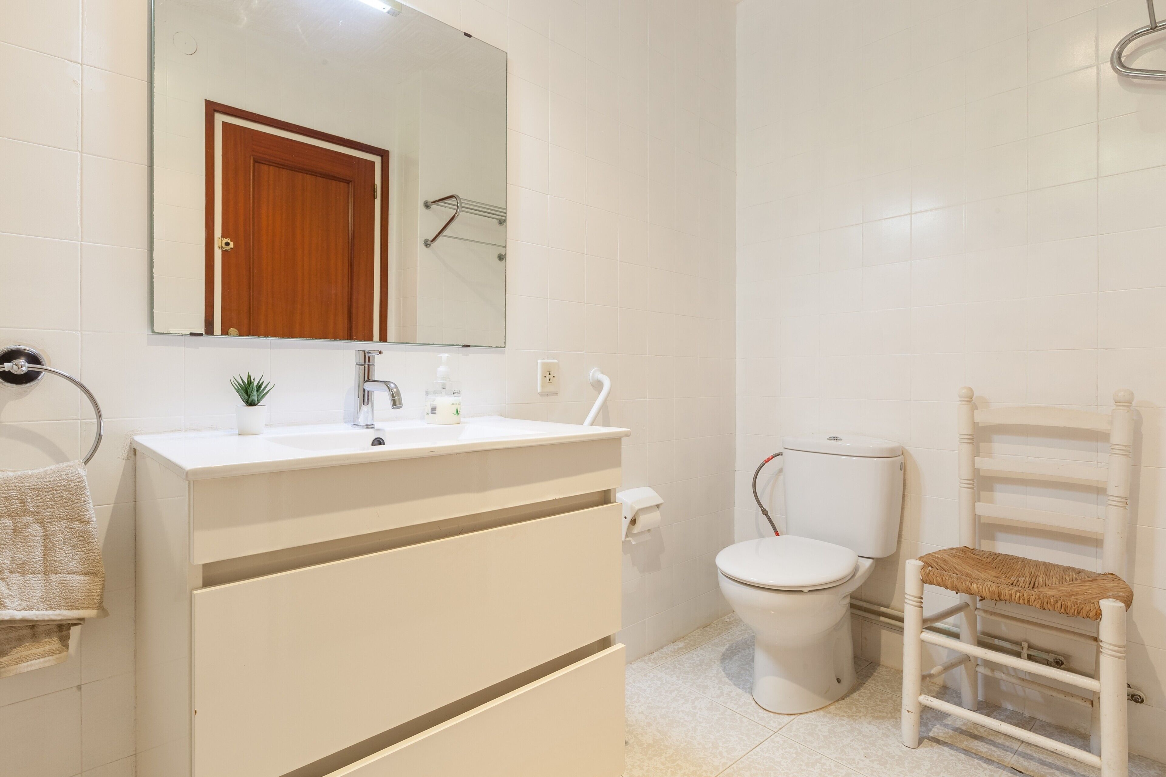 A bathroom with a white sink, mirror, toilet, and a wooden chair.