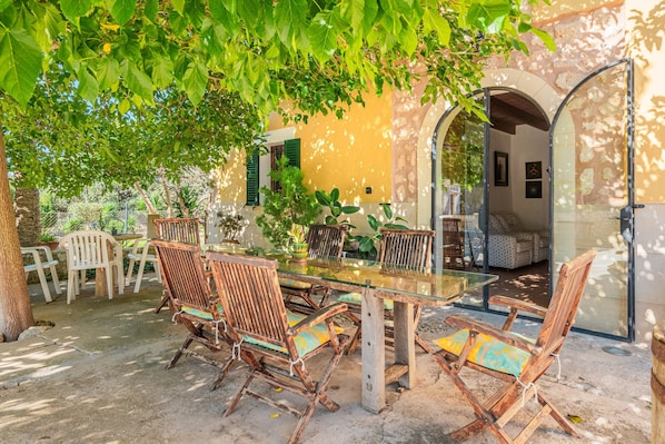 An outdoor dining area with a glass table and wooden chairs, surrounded by greenery and a yellow building with arches.