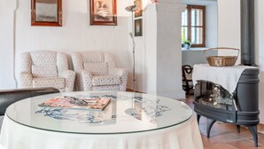 Ein Wohnzimmer mit rundem Glastisch, zwei gemusterten Sesseln, einem schwarzen Kaminofen und gerahmten Kunstwerken an der Wand.