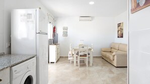 A compact living space with a washing machine, a dining table with chairs, a sofa, and a wall-mounted air conditioner.