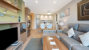 A modern living room with a sofa, a TV, a wooden coffee table, and a kitchen in the background.