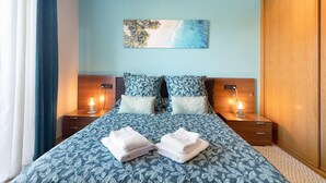 A bedroom with a bed, two bedside tables with lamps, a nightstand, a wooden wardrobe, and a painting on the wall.