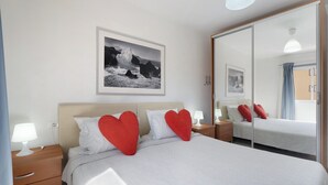 Ett hotellrum med en säng, sängbord, en spegel, en tavla på väggen och ett fönster med gardiner.