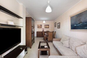A living room with a flat-screen TV, a sofa, a coffee table, a dining area with chairs and a table, and a painting on the wall.