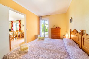 Une chambre à coucher comprenant un lit, une commode en bois, une table de chevet et une vue sur un coin repas, accessible par une porte ouverte.
