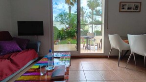 Una habitación con cama, televisión, mesa con un libro y una botella, y un ventanal corredero que da a un espacio exterior con sillas y una mesa.