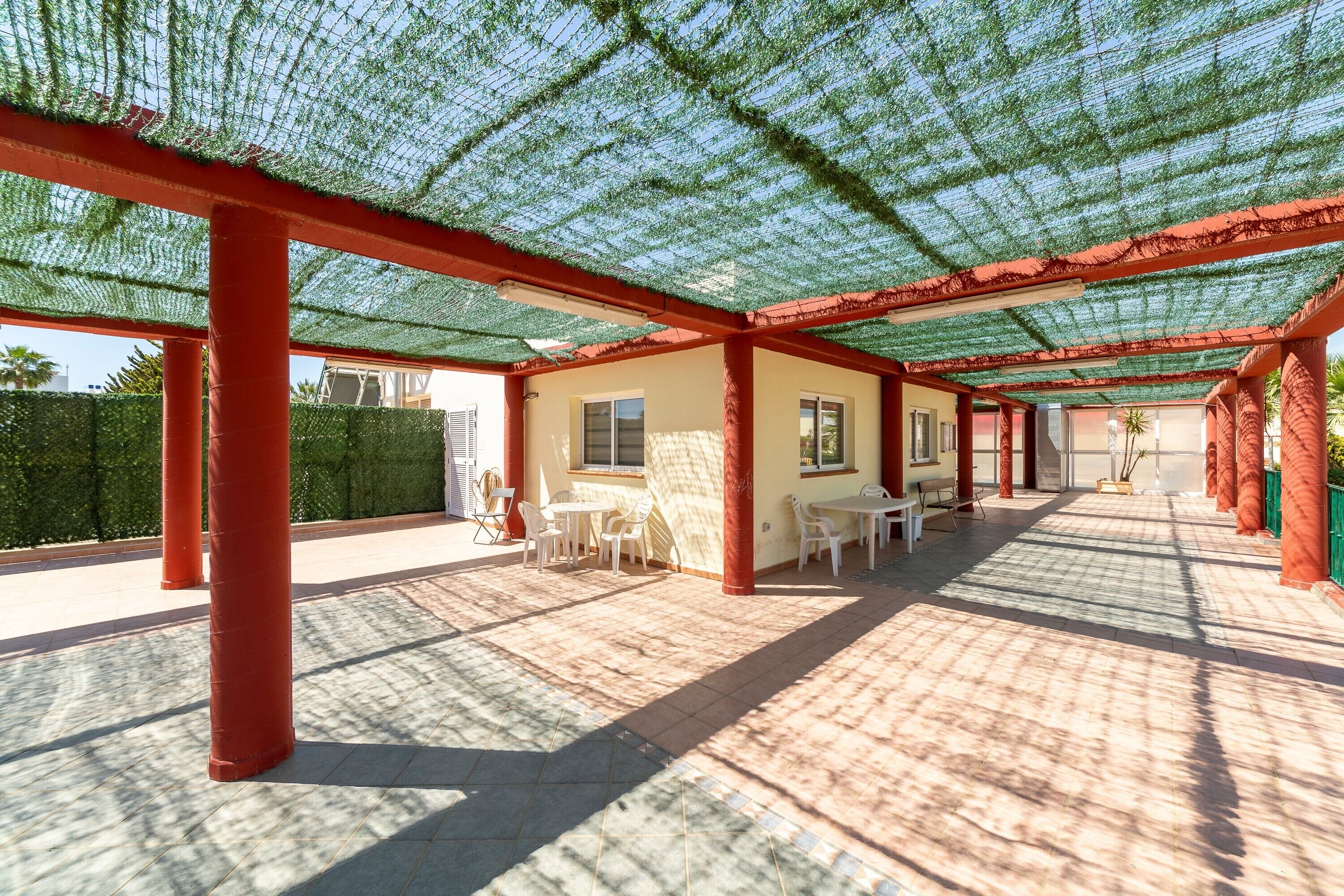 Un patio techado al aire libre con pilares rojos, suelo de baldosas y mesas y sillas blancas.