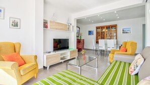 Un salon moderne doté d’un téléviseur à écran plat, de deux fauteuils jaunes, d’un canapé à rayures vertes et d’un coin repas avec un buffet en bois et une table blanche.