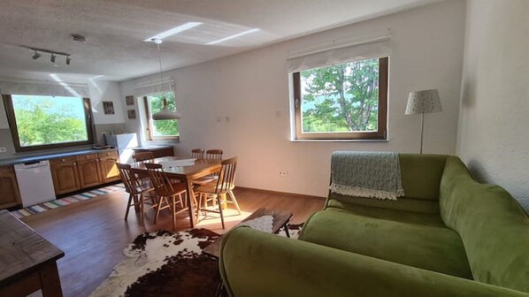 A spacious living area with a green sofa, a dining table with chairs, a kitchen area, and large windows with a view of greenery. - House on the edge of the Gacka Forest (Sinac)