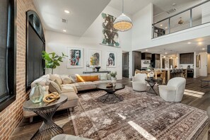 A spacious living area with a sofa, armchairs, a coffee table, and a rug. There is a kitchen in the background and a staircase leading to an upper level.