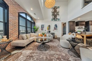 A spacious living area with a large window, a brick accent wall, and a modern kitchen in the background.