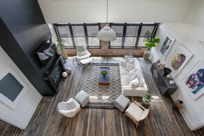 A spacious living area with a large sofa set, a coffee table, and a hanging light fixture. There are several chairs and a small side table. The room has large windows with a view of the outside. - Penthouse w Rooftop, Private Chef, Secure Parking (Atlanta)