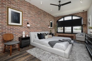 A modern bedroom with a brick wall, a large bed, a ceiling fan, and a large window.