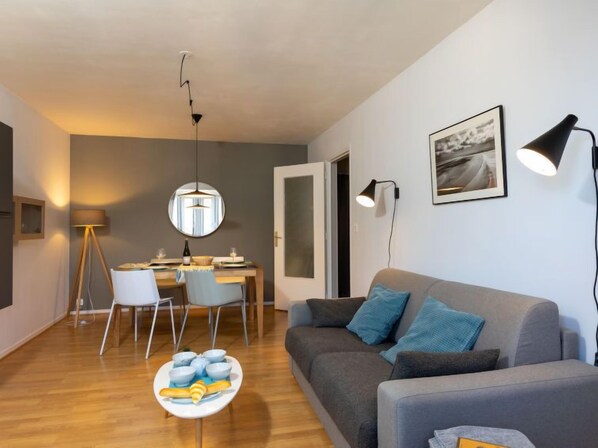 A modern living room with a grey sofa, a dining table with chairs, and a small round table with bowls and a baguette.