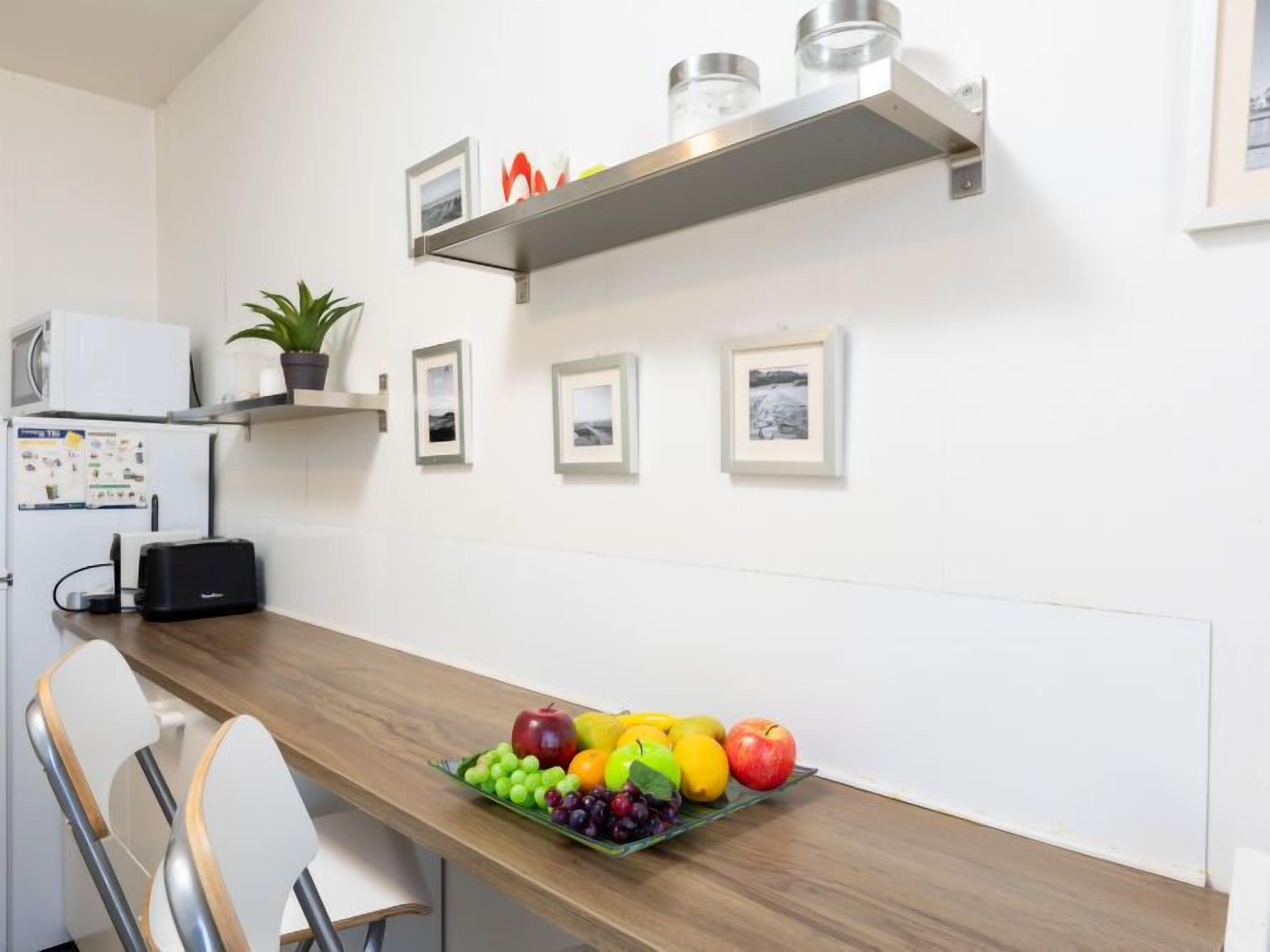 A modern kitchen with a wooden table, chairs, a fruit bowl, a microwave, a refrigerator, and framed pictures on the wall.