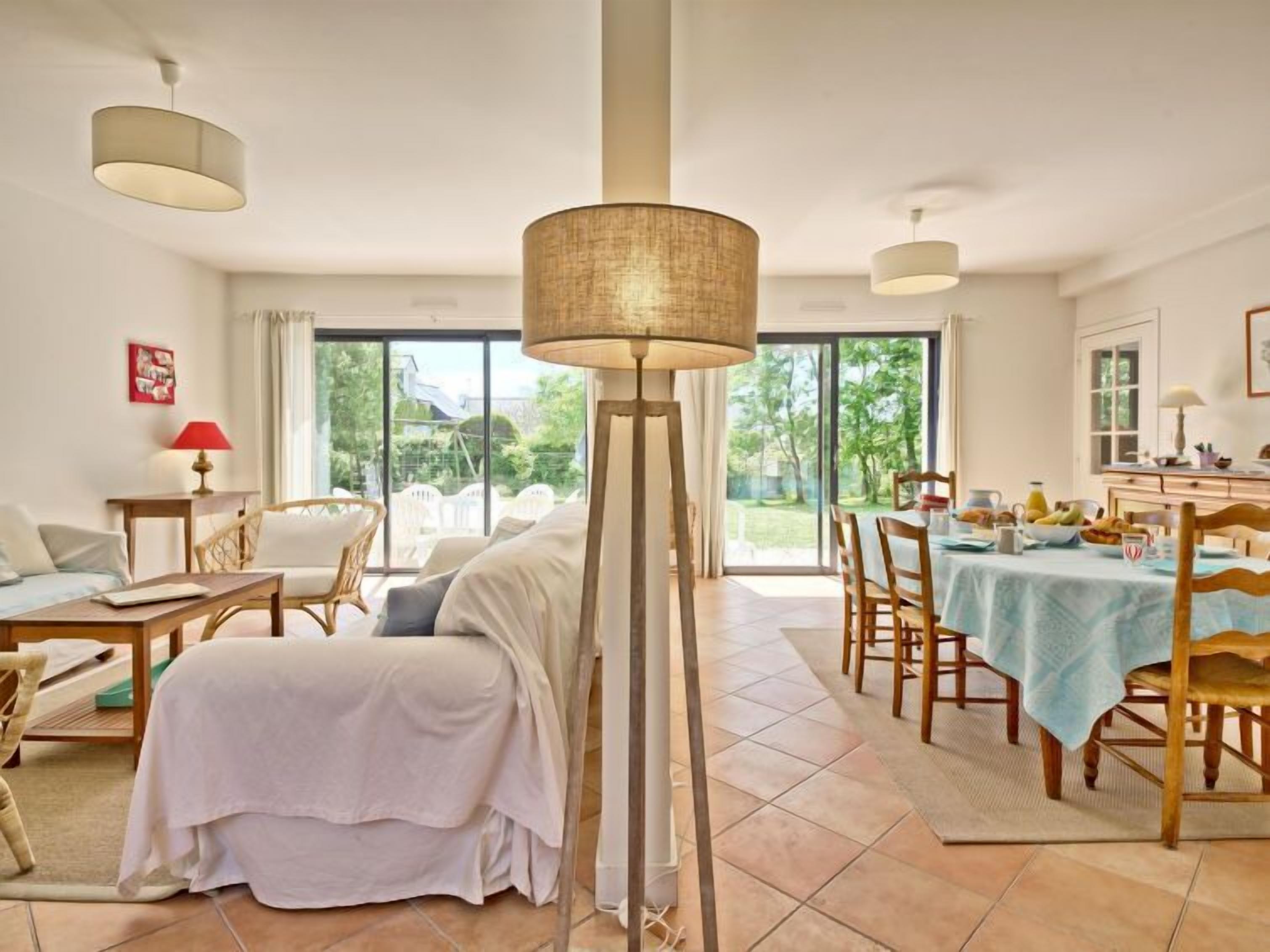Amplia sala de estar con sofá, mesa de comedor y sillas, ventanas grandes con vista a zonas verdes y una lámpara de pie alta.