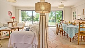 Amplia sala de estar con sofá, mesa de comedor y sillas, ventanas grandes con vista a zonas verdes y una lámpara de pie alta.