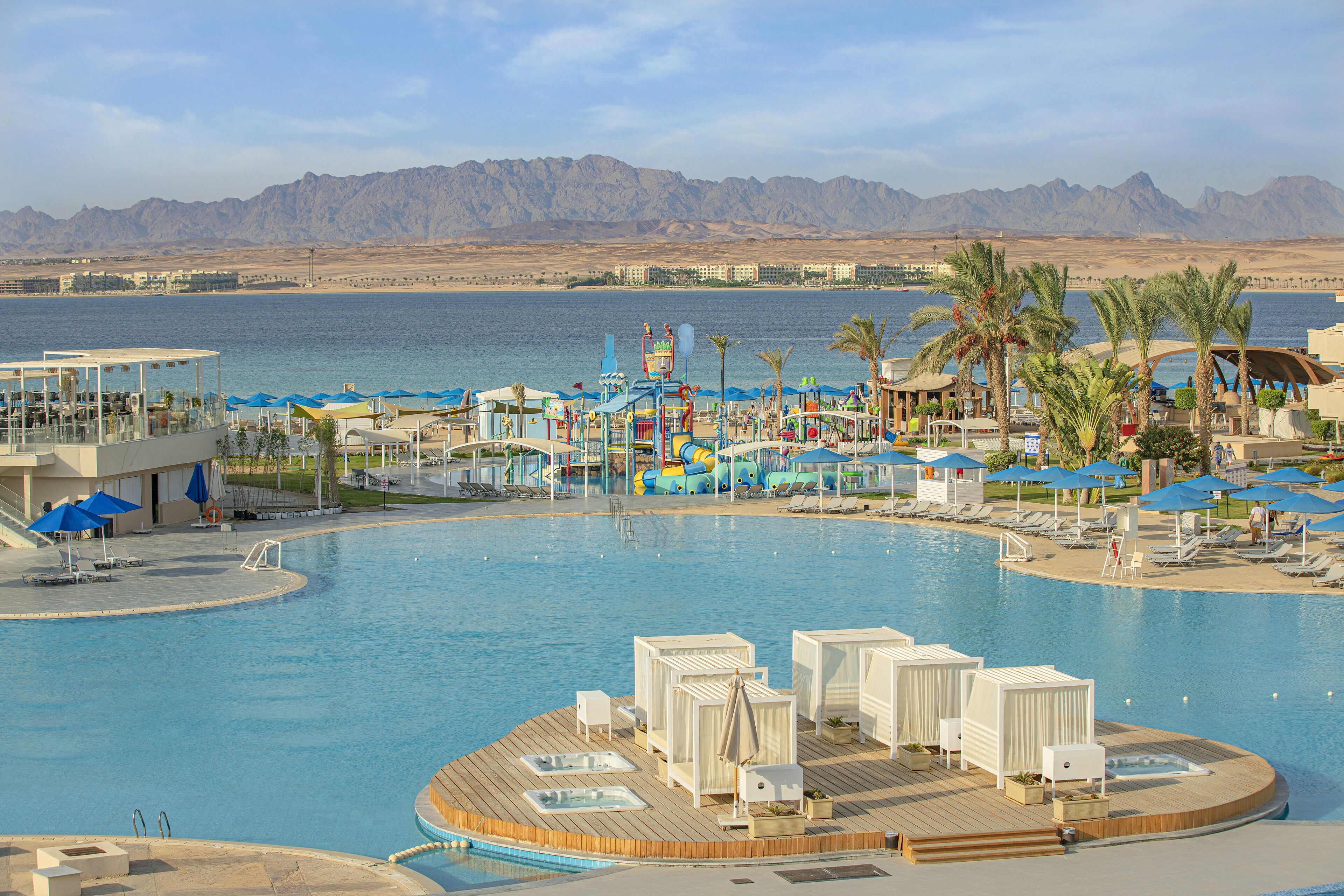 A resort pool area with a wooden deck, lounge chairs, umbrellas, and a view of the mountains.
