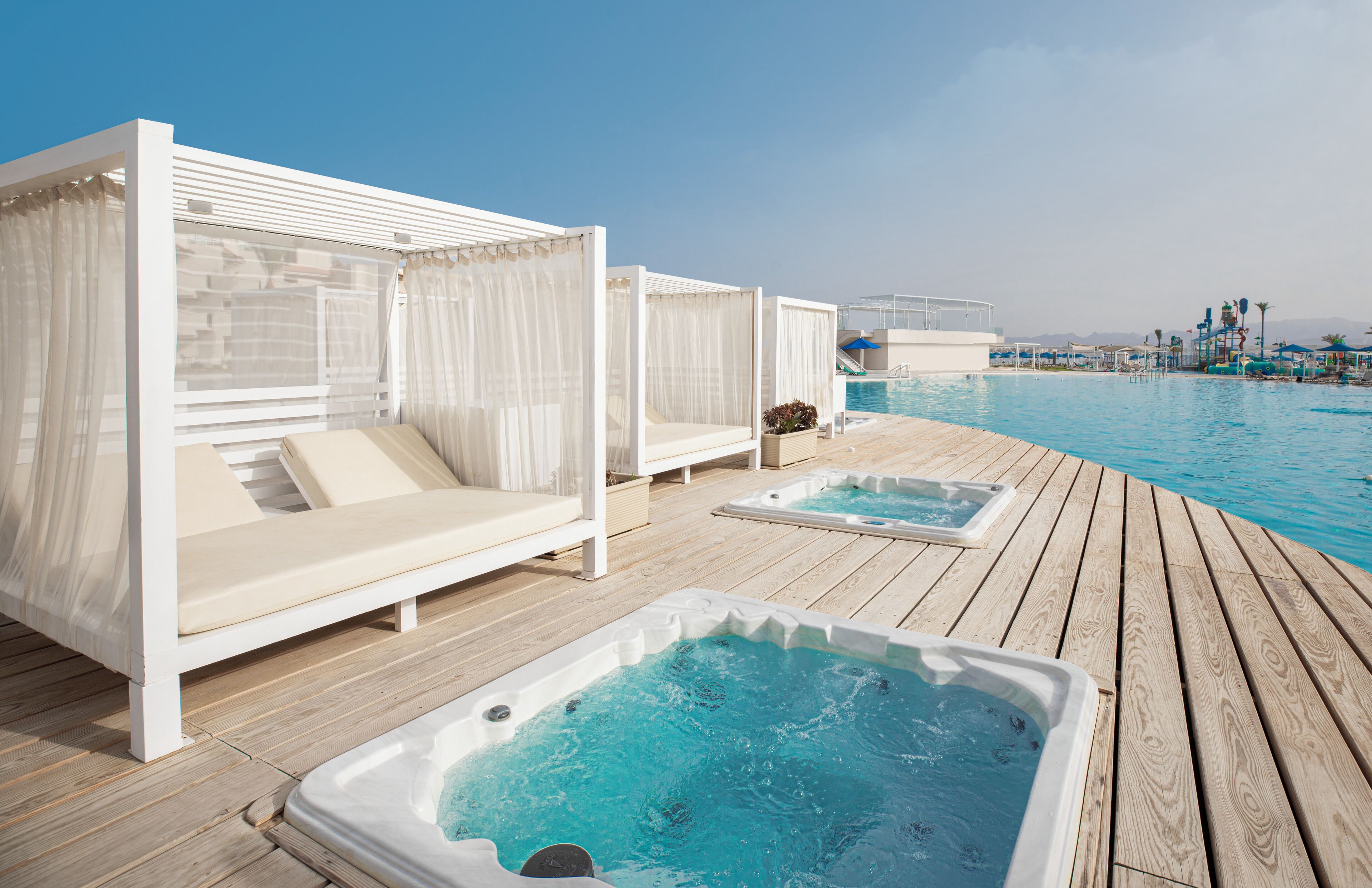 A wooden deck with two hot tubs and two white canopy beds with beige cushions.
