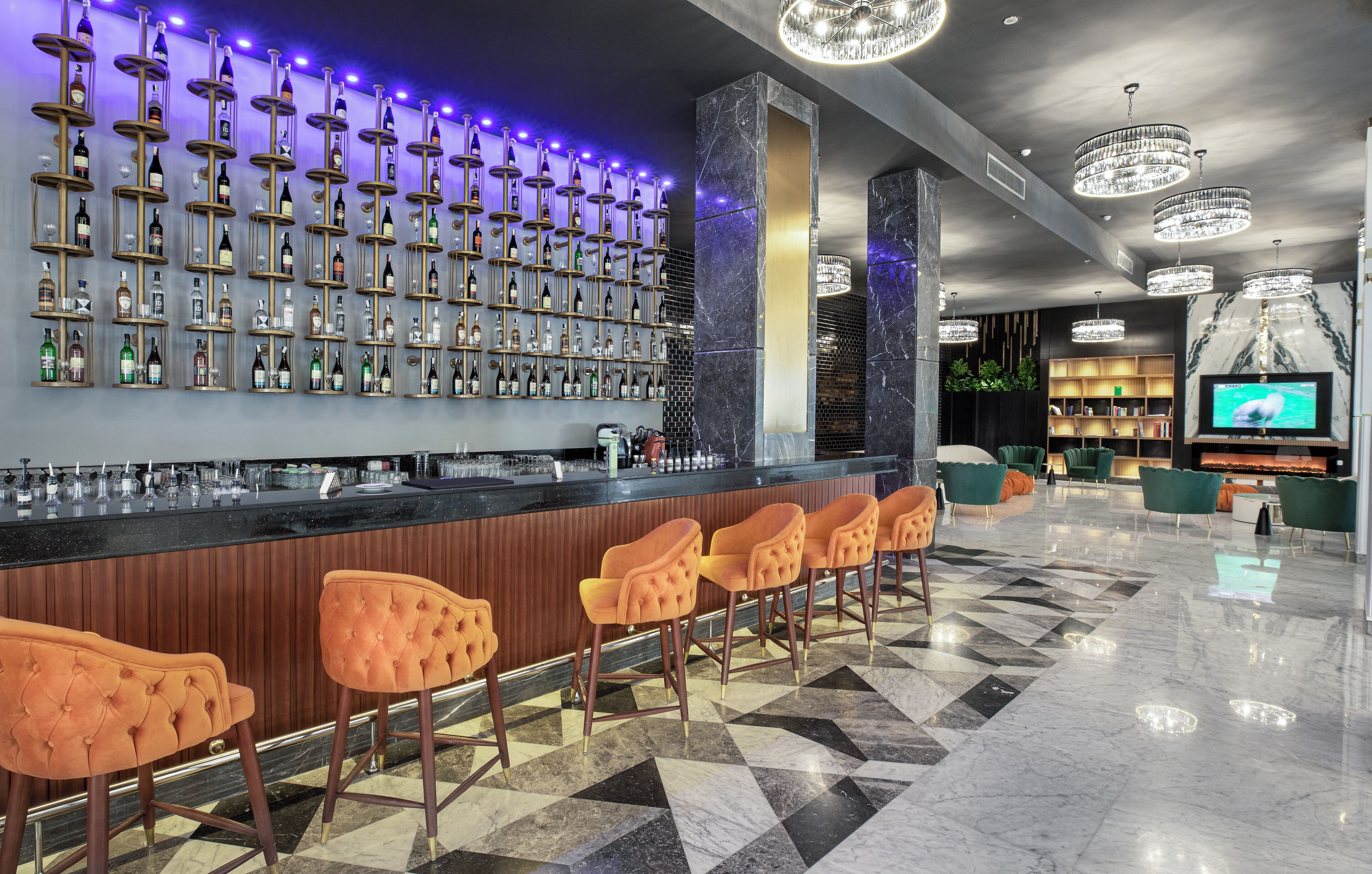A bar area with a long counter and multiple bar stools, a wall of neatly arranged bottles, and a television screen in the background.