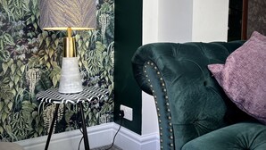 A room with a green upholstered sofa, a patterned wallpaper, a small side table with a lamp, and a purple pillow. - Beautiful Cottage in the heart of Lytham. (Lytham St.Annes)