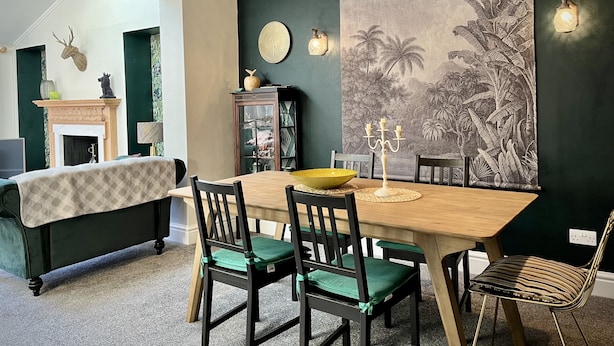 A dining area with a wooden table, green upholstered chairs, a candelabra, and a large tropical mural on the wall. - Beautiful Cottage in the heart of Lytham. (Lytham St.Annes)