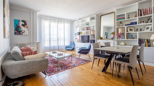 A modern living room with a sofa, coffee table, and chairs, a dining area with a table and chairs, and a bookshelf filled with books and decorative items. - Dreaming in the Daytime, by Veeve (Paris)