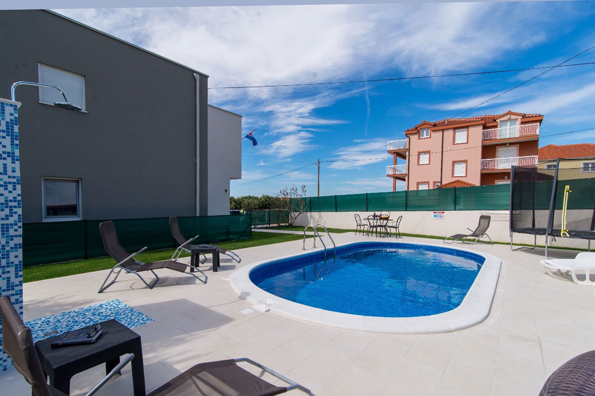 A swimming pool with lounge chairs and a table, surrounded by a fenced area with a building in the background.