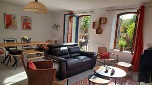 Ein modernes Wohnzimmer mit einem schwarzen Ledersofa, Korbstühlen, einem Esstisch und einem großen Fenster mit Vorhängen.