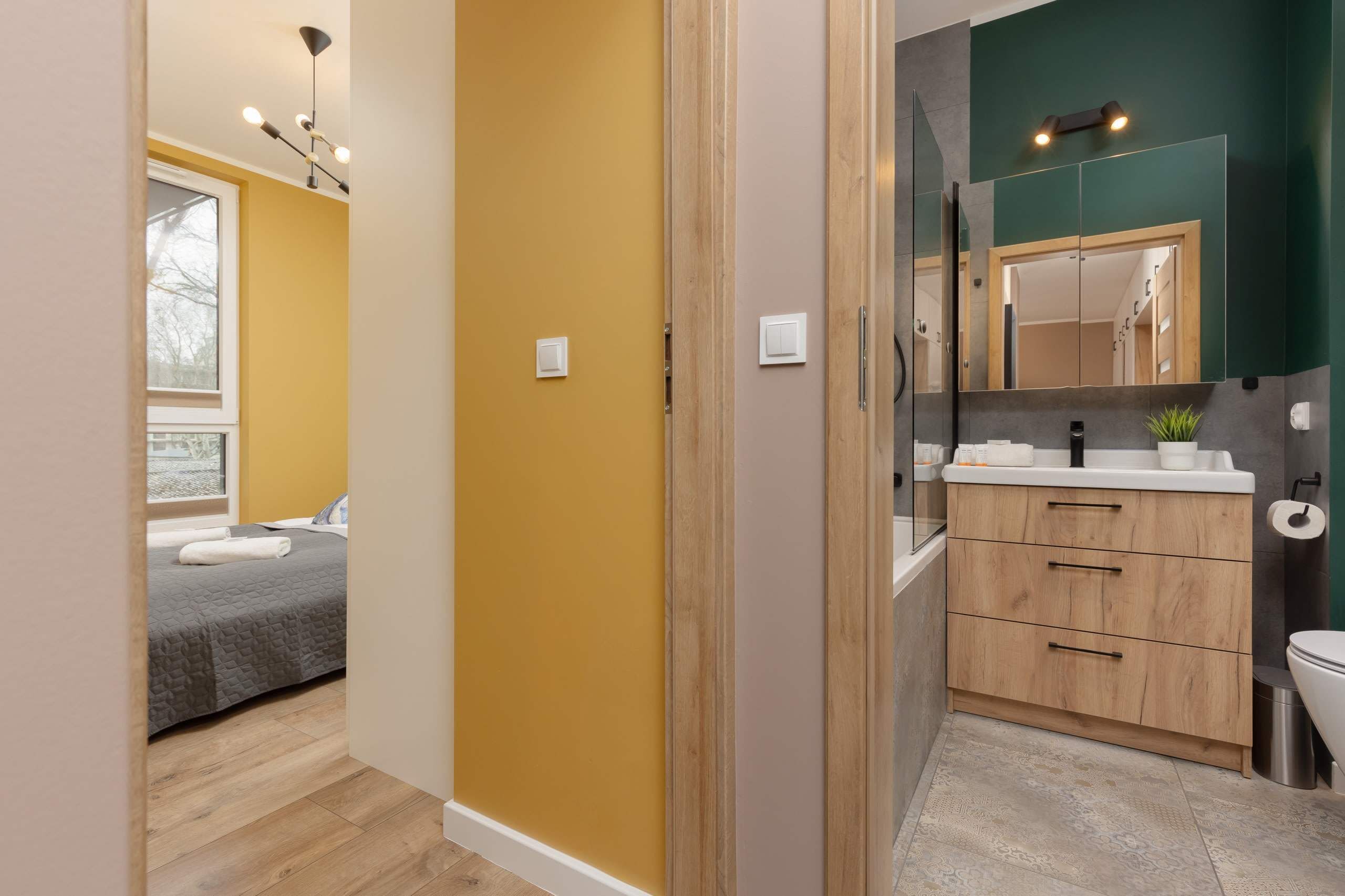 A modern hotel room with a bed, a bathroom with a mirror, sink, and cabinets.