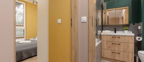 A modern hotel room with a bed, a bathroom with a mirror, sink, and cabinets.