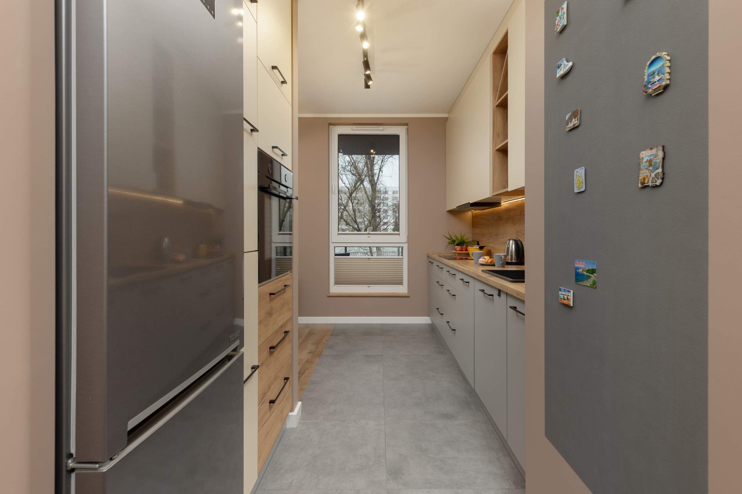 A narrow kitchen with a stainless steel refrigerator, wooden cabinets, and a window with a view of trees.