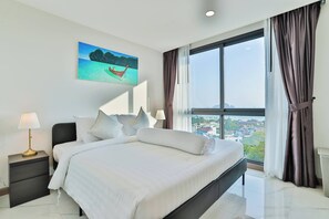 A modern bedroom with a large bed, a nightstand with a lamp, a painting of a tropical scene, and a large window with curtains.